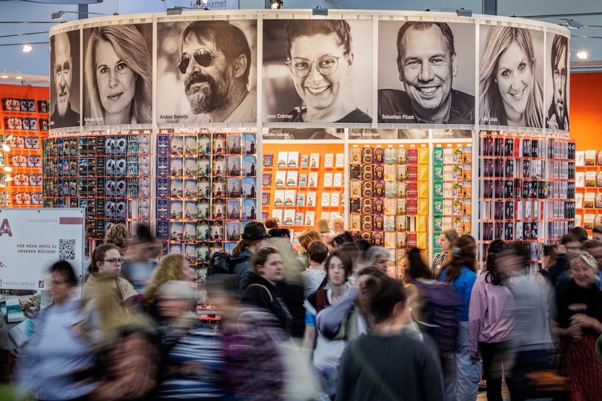 تصویر غرفه‌ای از نمایشگاه کتاب لایپزیگ را نشان می‌دهد که بر سردر آن تصاویری از نویسندگان دیده می‌شود و در مقابل آن جمعیت در حال رفت‌وآمدند.
