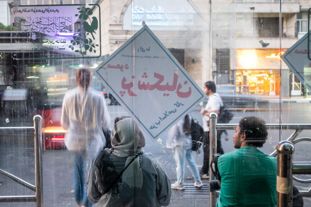 یک مرد و زن در ایستگاه اتوبوس تهران نشسته اند. عکس از پشت سر گرفته شده است. پوستر پزشکیان هم دیده میشه
