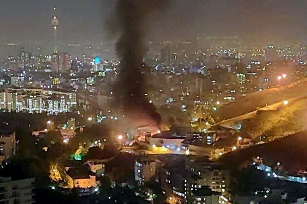 تصویر نمای دور از زندان اوین در شب آتش‌سوزی است. از محوطه زندان دود به آسمان برخاسته و در پسزمینه تهران و برج میلاد دیده می‌شود.