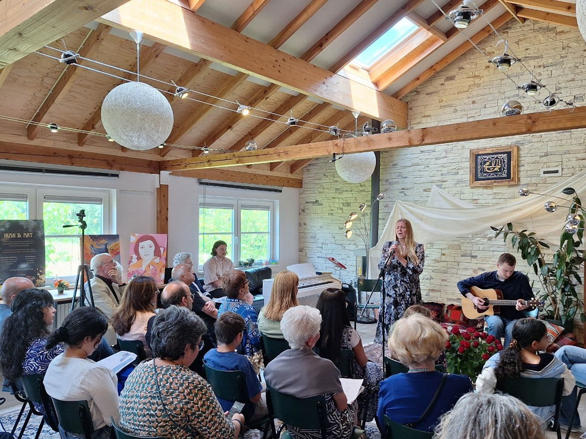 اتاقی با سقف زیرشیروانی چوبی، جمعیت در اتاق نشسته‌اند و جلوی اتاق یک زن در خواندن و یک مرد در حال گیتار زدن است