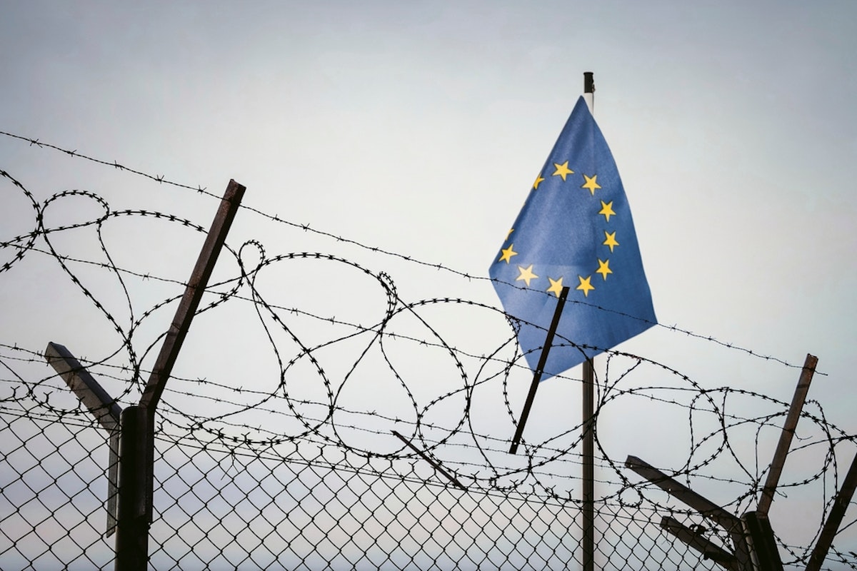 پرچم اتحادیه اروپا که برفراز دیواری از سیم خاردارها و فنس افراشته شده است.