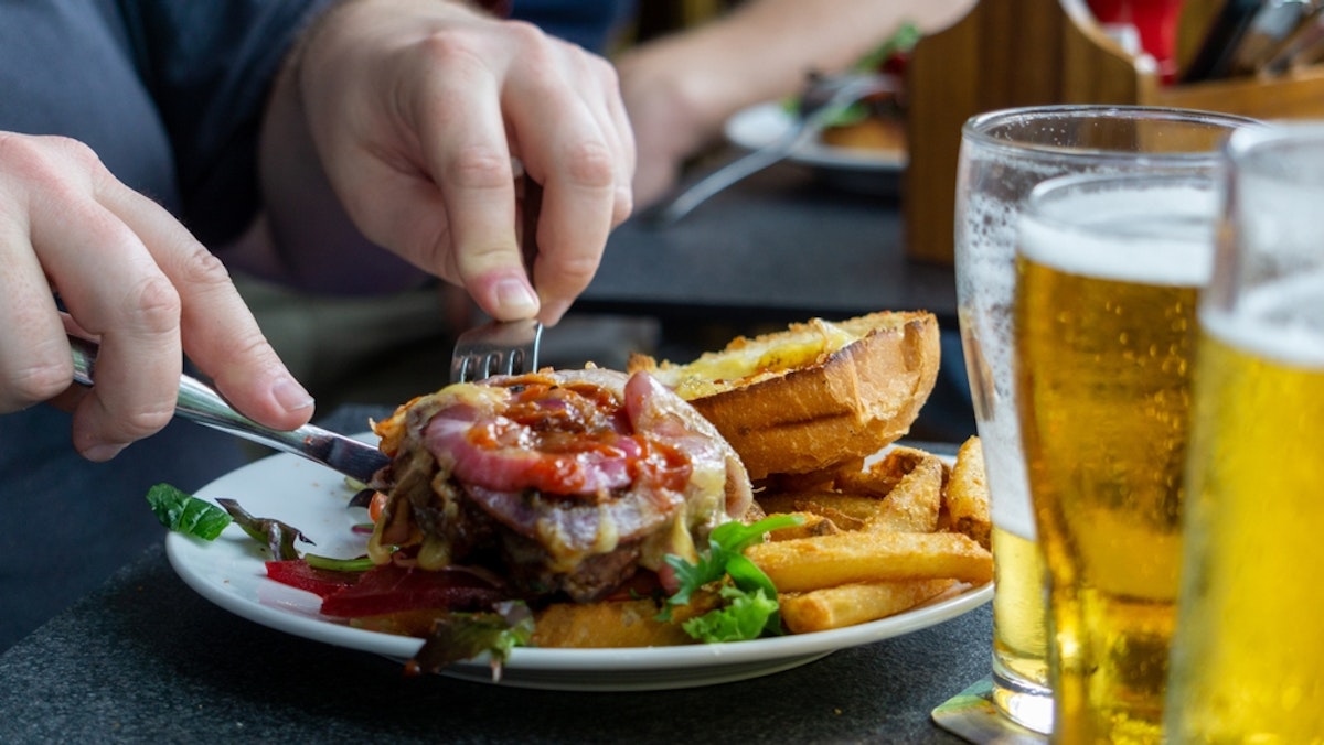 عکس دو دست در حال بریدن همبرگر، همراه با سیب‌زمینی سرخ‌کرده و لیوان‌های آبجو