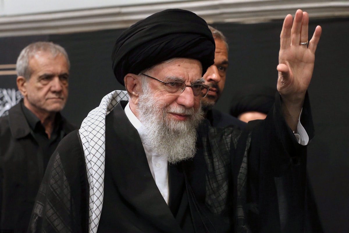 علی خامنه‌ای در حال دست تکان دادن برای هوادارانش. مسعود پزشکیان در پسی زمینه دیده می‌شود