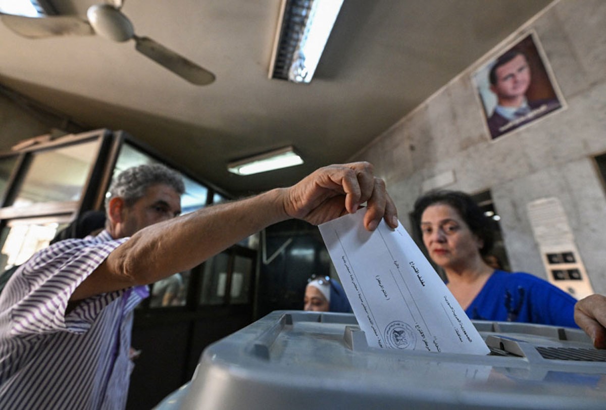 مردی در کنار یک زن دارد رأی خود را به صندوق می‌ریزد. در پسزمینه‌ی تصویر عکس بشار اسد، رهبر سوریه بر دیوار دیده می‌شود.