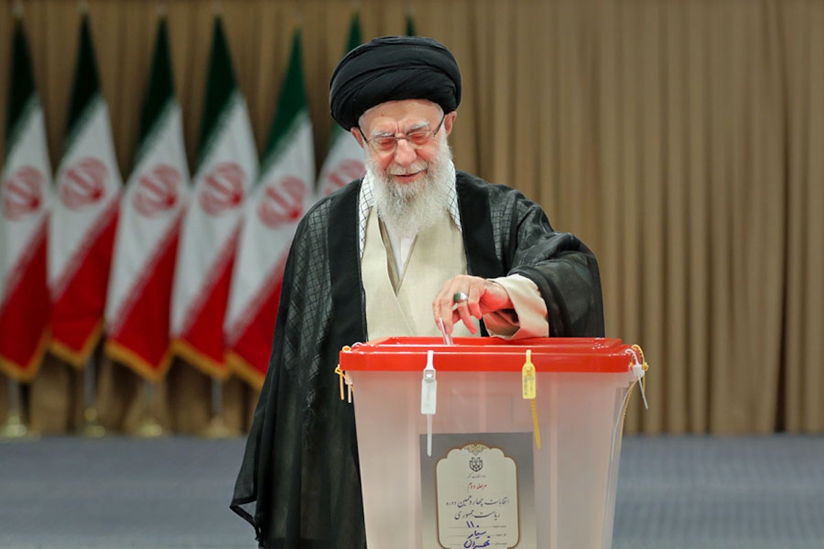 علی خامنه‌ای، رهبر جمهوری اسلامی برگه رأی را در صندوق رأی‌گیری می‌اندازد. پشت سر او چندین پرچم جمهوری اسلامی دیده می‌شود.